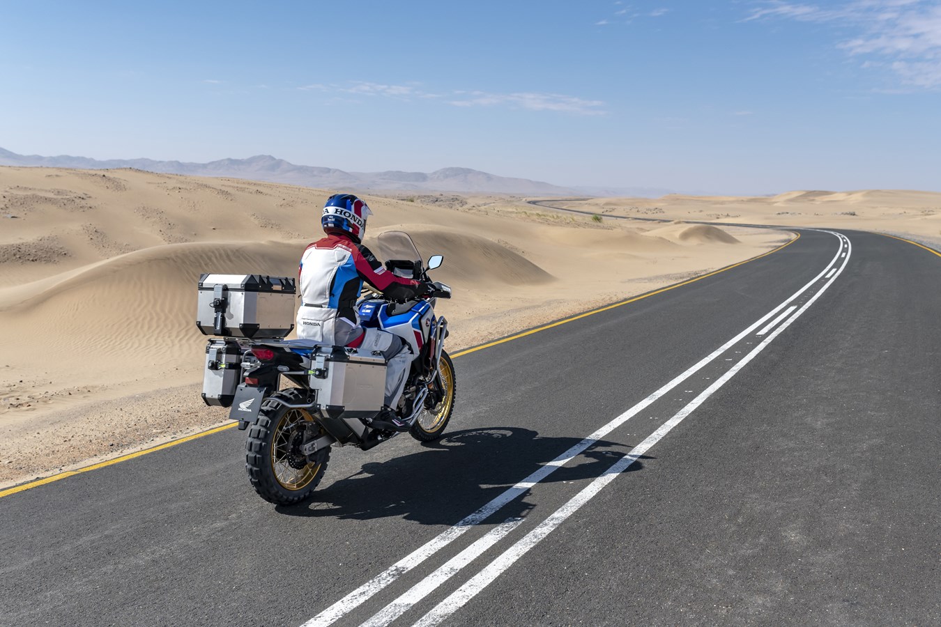 Piloto em Honda Africa Twin viajando de moto em estrada com areia em volta