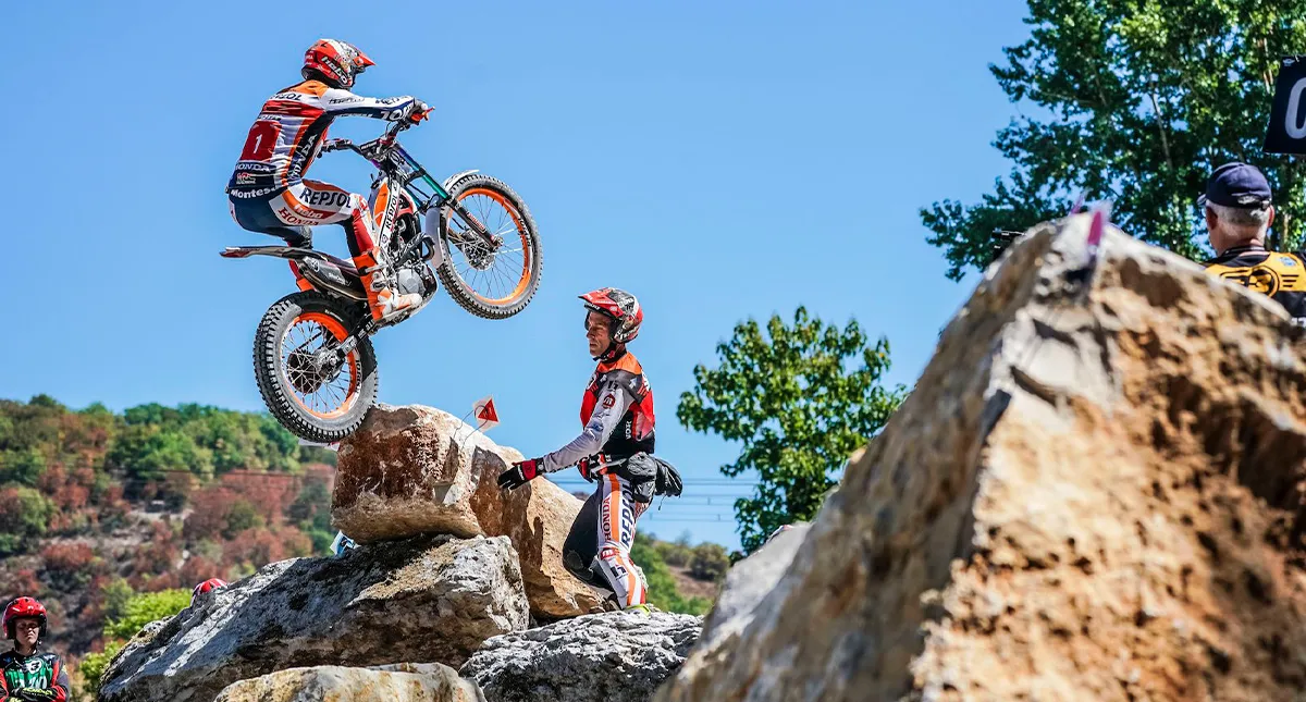 Piloto da Honda Toni Pulando de uma Pedra para Outra e Piloto Vestido de Laranja Olhando
