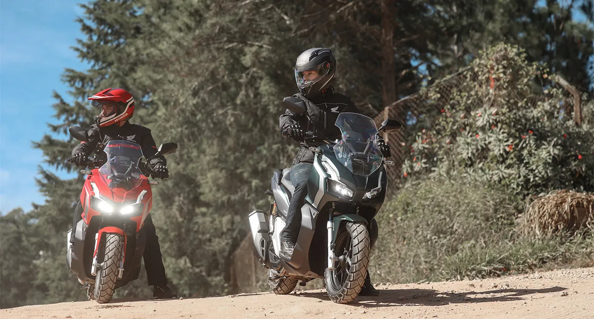 Mulher em Honda ADV preta parada olhando estrada de terra e homem em honda adv vermelha atrás