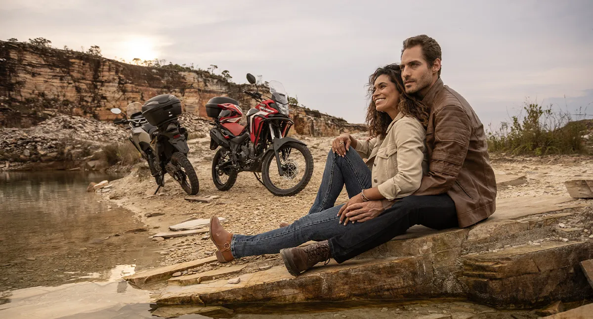 Casal sentado em meio a natureza admirando a paisagem com suas motocicletas Honda Sahara ao fundo
