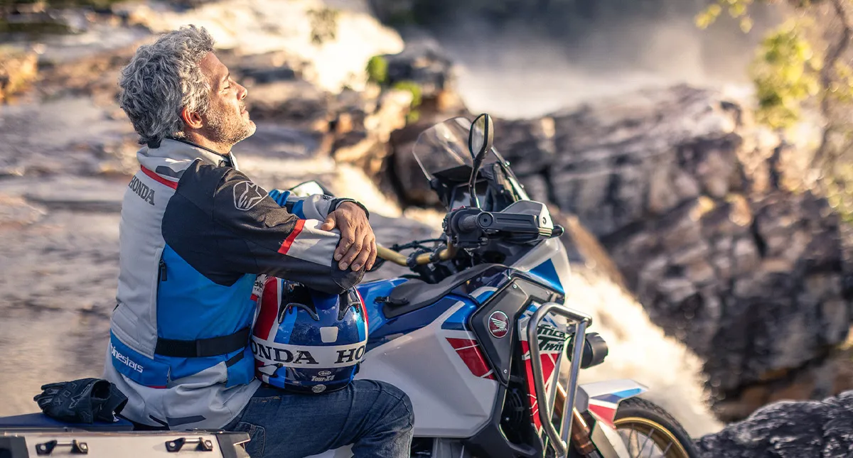 Homem sentado vestindo jaqueta de proteção Honda, com capacete no colo ao lado de sua CRF 1100L a Africa Twin admirando a paisagem