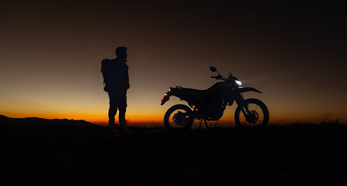 Homem olhando Moto Honda XR 300L Tornado com farol aceso e pôr do sol
